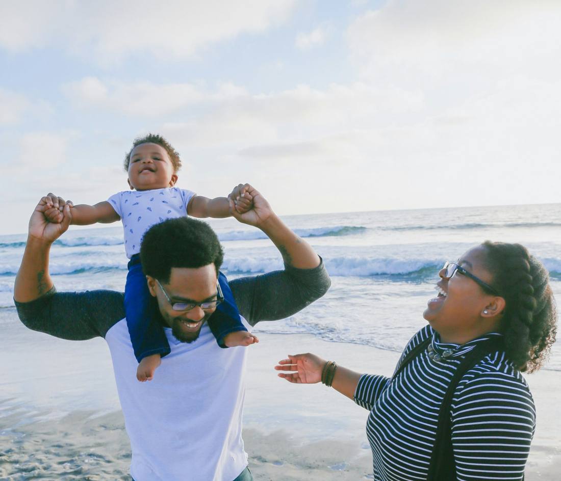 A dad lifts his laughing baby on his shoulders at the beach as mom laughs along